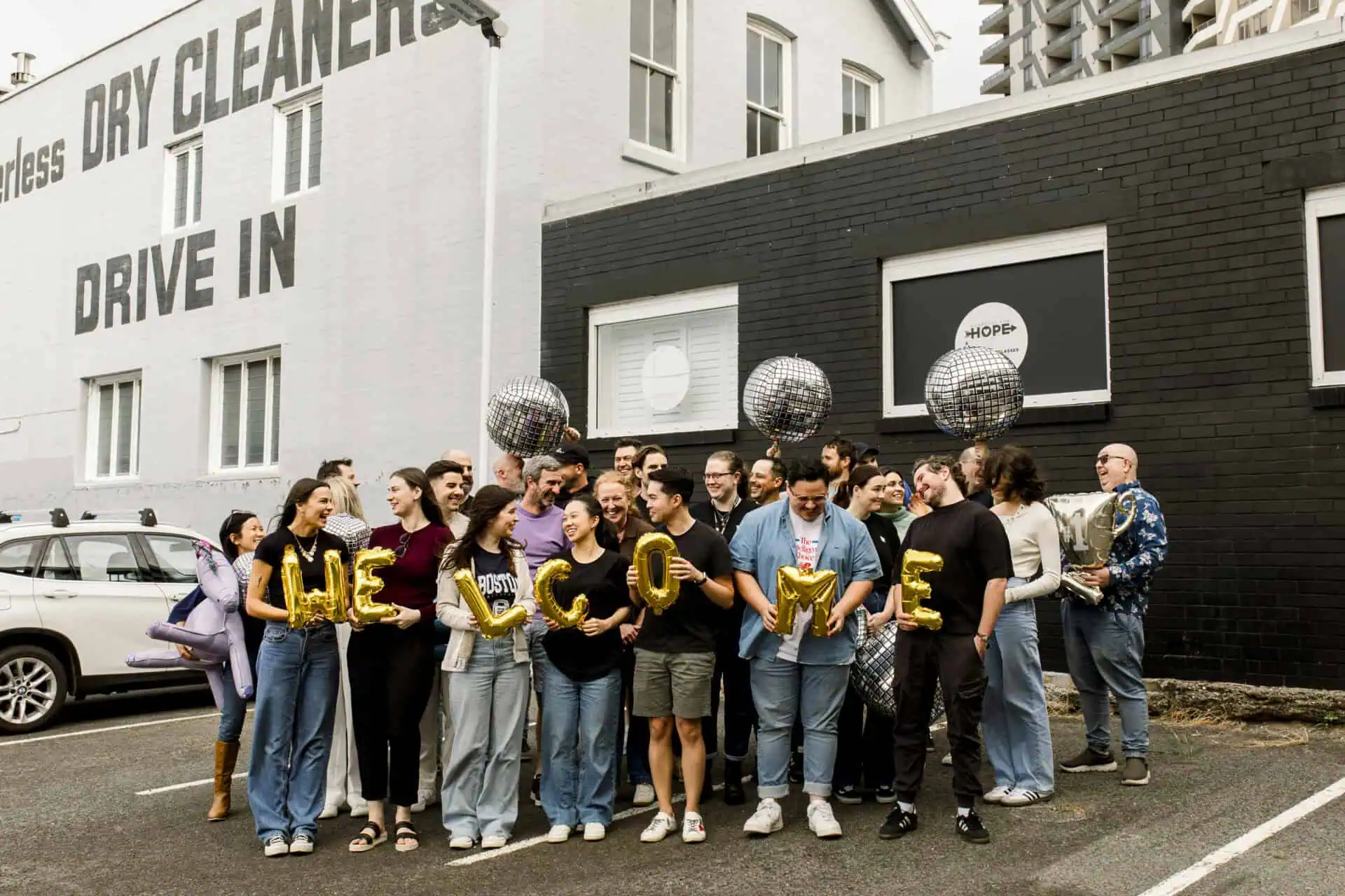 Excite Media team holding gold foil balloons in a car park that read 'welcome'