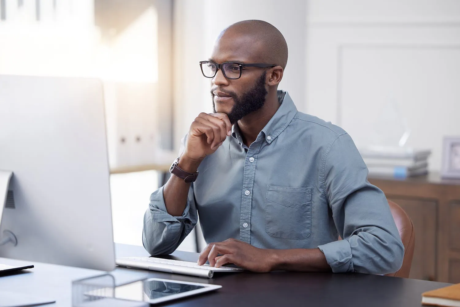 black man with glasses holds his beard white looking at the computer